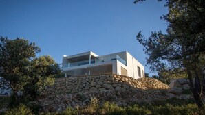 A modern two-story house with a stone foundation, surrounded by trees and a clear sky.