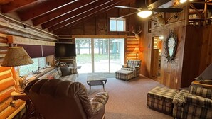 A log cabin interior with a cozy seating area, a television, and a large window overlooking a grassy area.