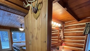 A log cabin interior with a wooden wall, a mounted deer head, a bathroom with a sink and mirror, and a bed with a green rug.