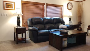 A living room with a leather sofa, a wooden coffee table, two side tables with lamps, a clock on the wall, and a vase with dried plants.