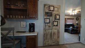 A kitchen with a coffee maker, a wooden cabinet with glassware, and a door with framed pictures.