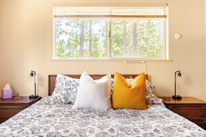 A bed with patterned bedding, a nightstand with a lamp, and a window with blinds. - Spacious Chalet with Game Loft & Pool Access (Arnold)