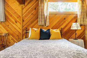 Ein Zimmer mit Holzwänden, einem Bett, Vorhängen, einem Tisch mit einer Lampe und einem Fenster mit Blick auf Bäume.