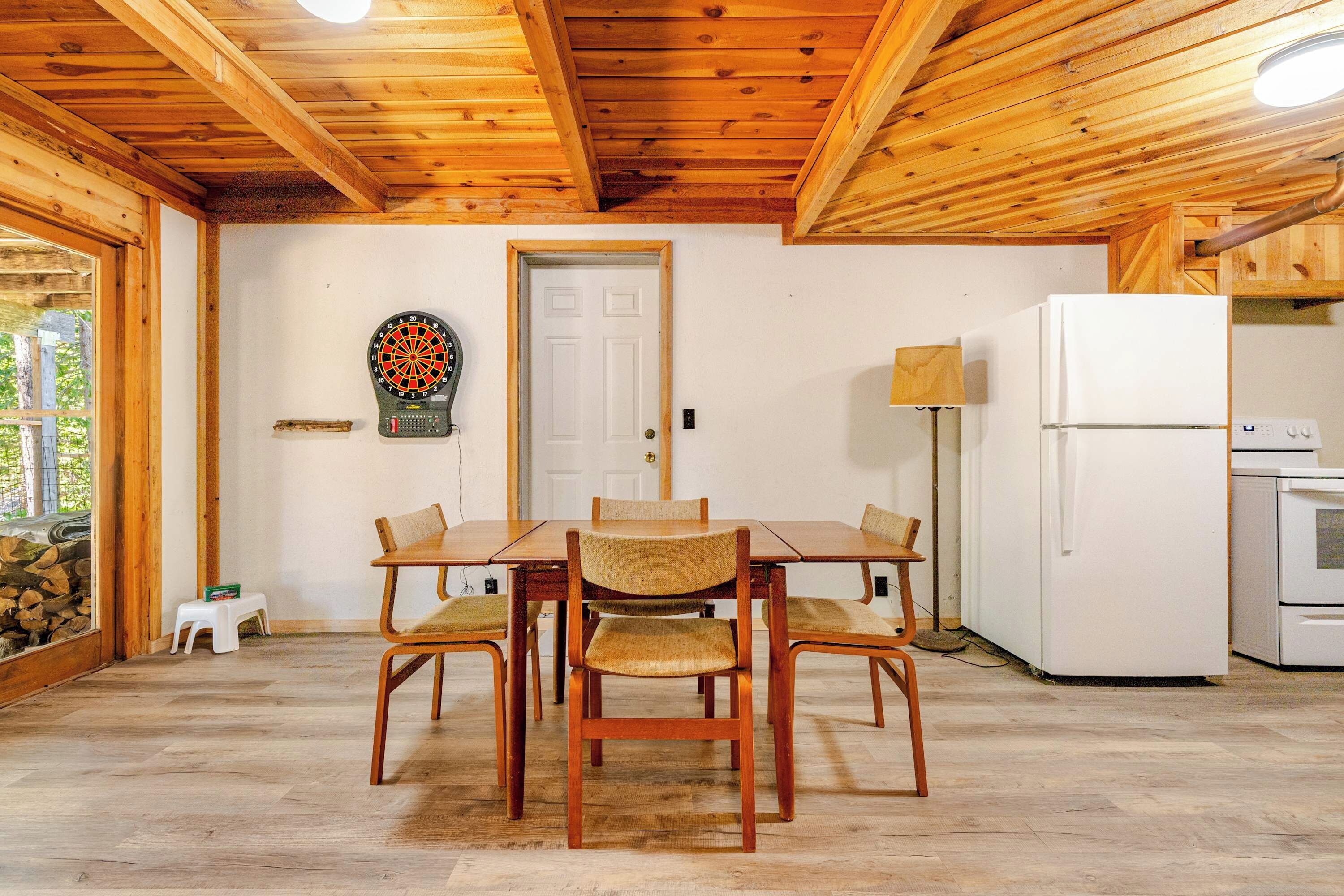 A wooden-paneled room with a dining table and chairs, a dartboard, a white door, and a refrigerator.