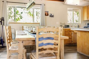 A dining area with a wooden table and chairs, a kitchen in the background, and large windows with a view of trees. - Spacious Chalet with Game Loft & Pool Access (Arnold)