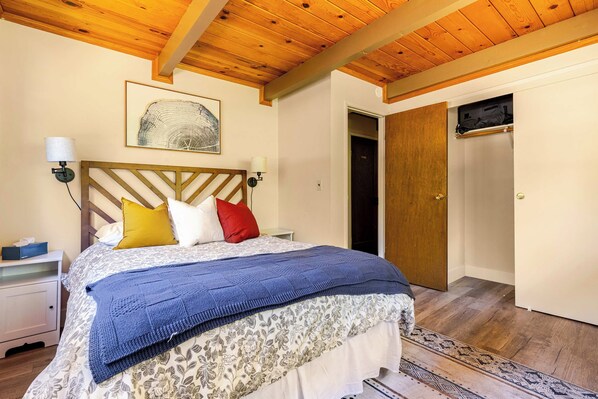 A bedroom with a wooden ceiling, a bed with a patterned bedspread, two bedside lamps, a framed artwork on the wall, and a door to the right. - Spacious Chalet with Game Loft & Pool Access (Arnold)