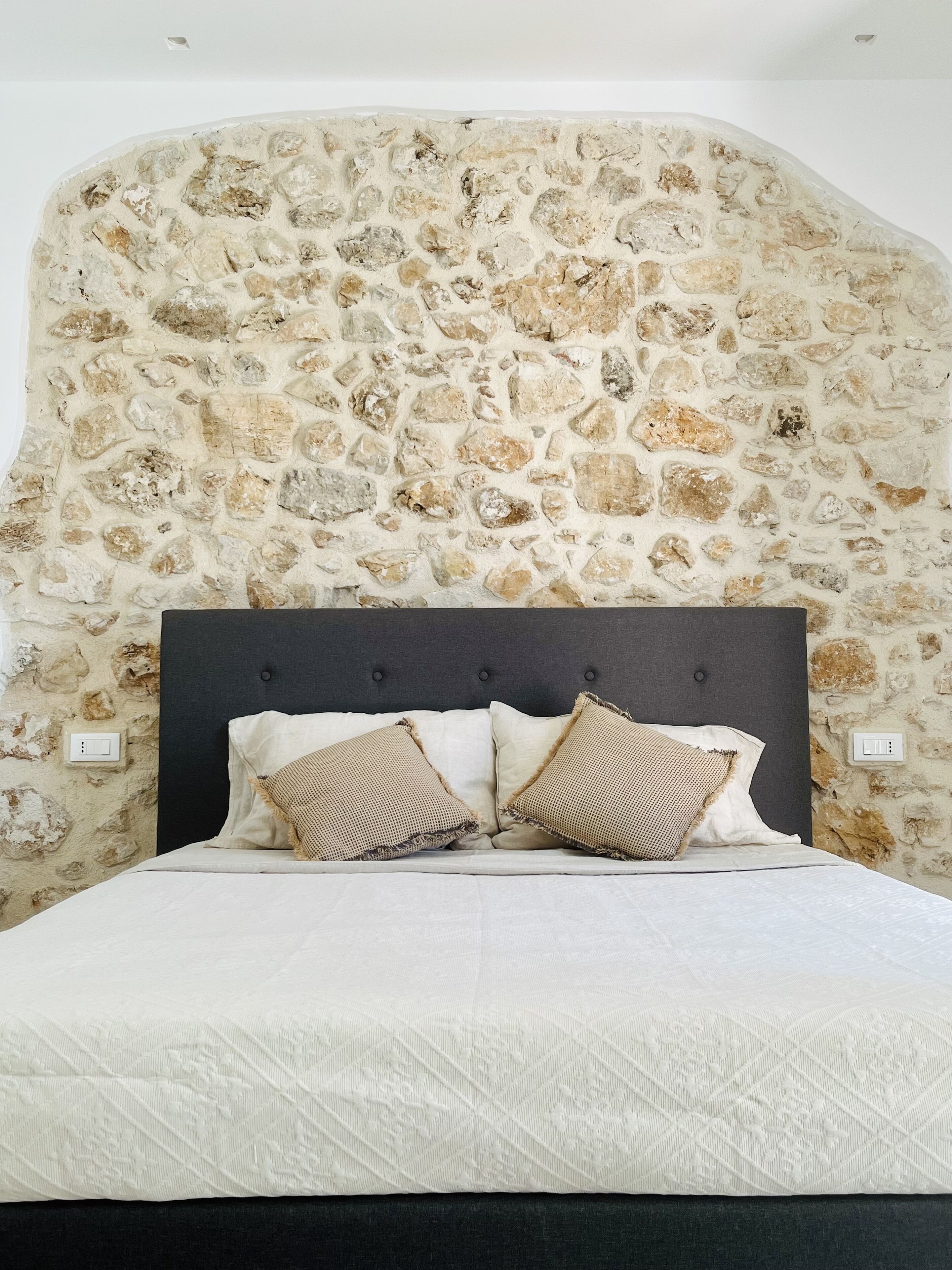 A bedroom with a stone wall, a dark headboard, and two patterned pillows.