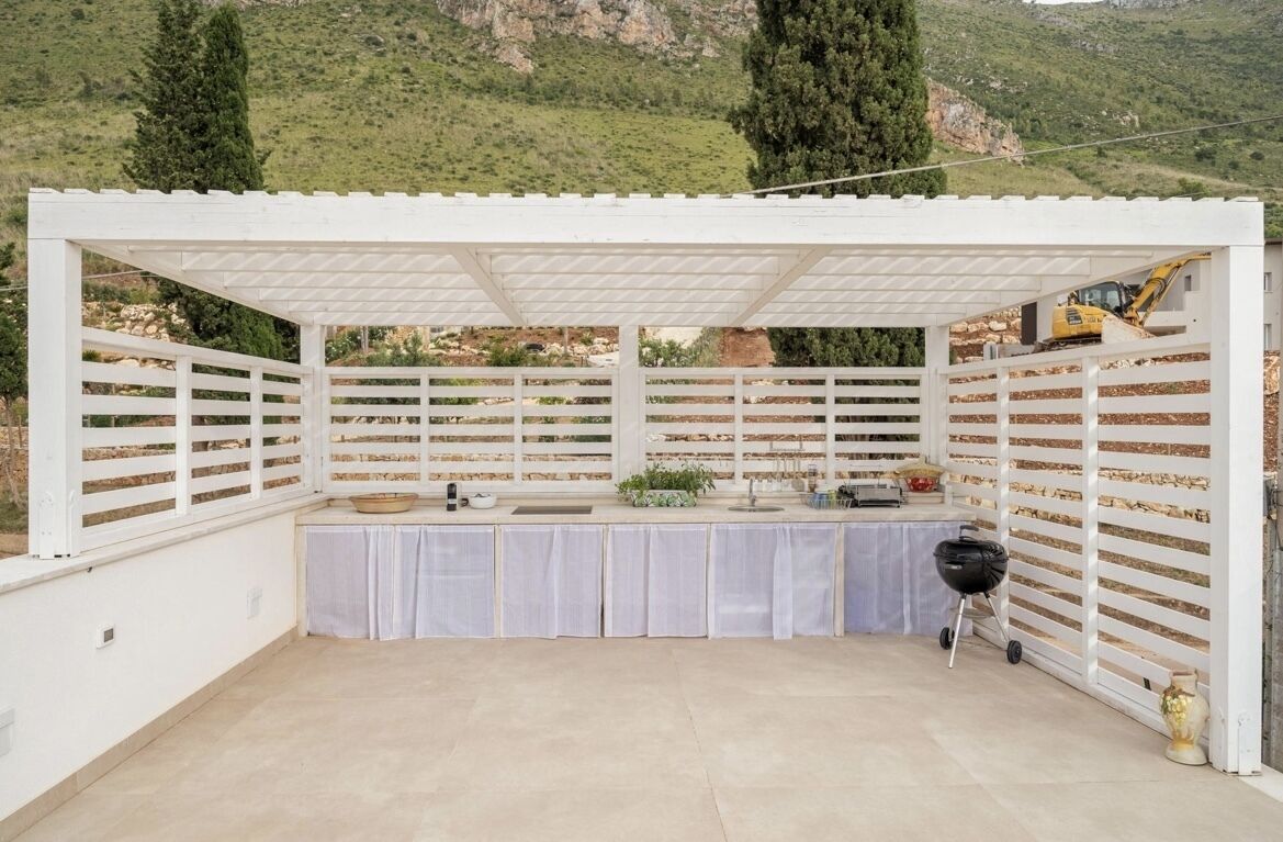 A covered outdoor area with a white pergola, a barbecue grill, and a table set up for dining.