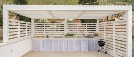 A covered outdoor area with a white pergola, a barbecue grill, and a table set up for dining.