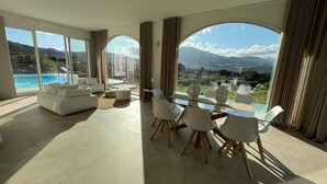 A spacious living room with a large glass table and white chairs, a sofa, and a view of a pool and mountains. - Curina Luxury Villa - design pool villa in Scopello (Castellammare del Golfo)