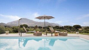 A pool with lounge chairs and an umbrella, surrounded by greenery and mountains in the background. - Curina Luxury Villa - design pool villa in Scopello (Castellammare del Golfo)