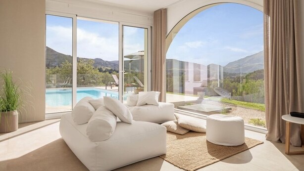 A modern living room with a large sofa, a small ottoman, and a view of a swimming pool and mountains. - Curina Luxury Villa - design pool villa in Scopello (Castellammare del Golfo)