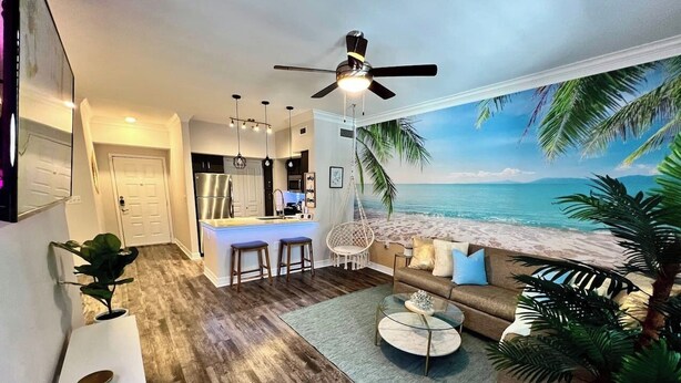 Una sala de estar con un mural de playa, piso de madera, ventilador de techo, un sofá, una mesa de centro y una zona de cocina con refrigerador y taburetes de bar.