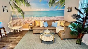 Una sala de estar con un mural de playa, una silla colgante, un sofá con cojines y una mesa de centro.
