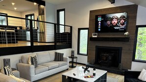 Una sala de estar moderna con un sofá gris, almohadas negras y blancas, una mesa de centro con una botella de vino y un televisor mostrando 'Netflix'.