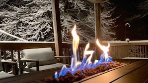 Una fogata en una terraza de madera con árboles nevados al fondo.