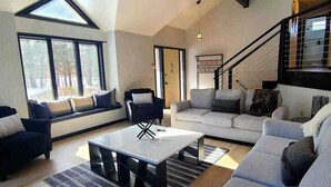 Una sala de estar moderna con sofá, sillones y una mesa de centro. Se ve una escalera con barandilla que sube a un nivel superior.