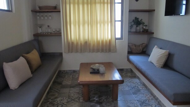 Ein Wohnzimmer mit zwei Sofas, einem Couchtisch, gemusterten Bodenfliesen, einem Fenster mit Vorhängen und Regalen mit Gegenständen.