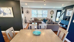 Uma área de refeições com mesa e cadeiras de madeira, um compasso decorativo na parede e uma janela com persianas.