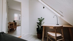 A room with a sloped ceiling, a desk with a chair, a potted plant, and a view into another room with a dining table and chairs. - Bergen Beds - The Penthouse (Bergen)