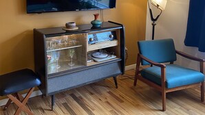Una sala de estar con un televisor de pantalla plana montado en la pared, un mueble de entretenimiento de estilo vintage con puertas de cristal, un sillón azul y una lámpara de pie.