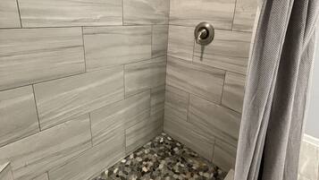 A shower with a stone mosaic floor, grey tiles on the walls, and a window with a blue view.