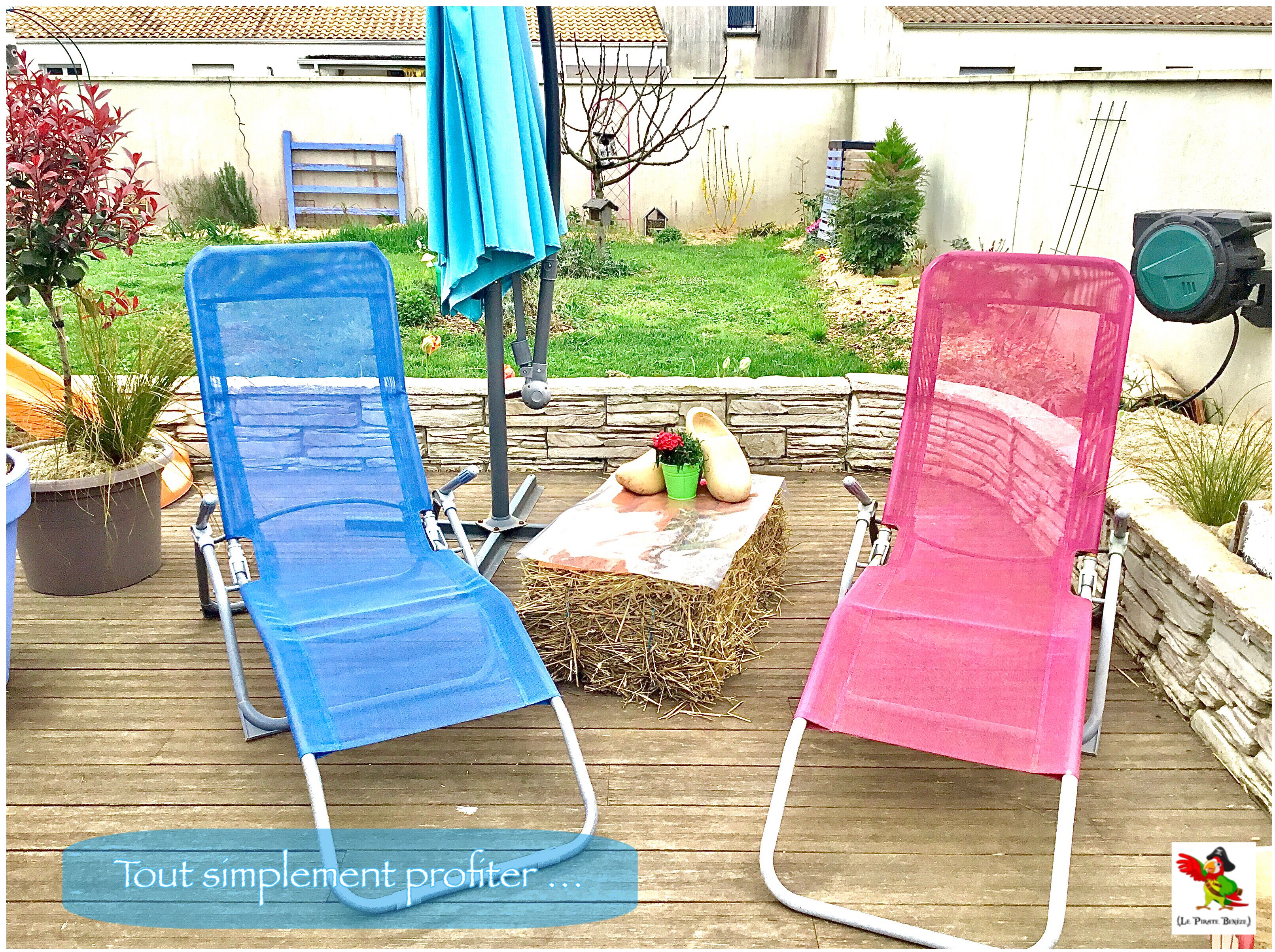Deux fauteuils de jardin sont placés sur une terrasse en bois, adossés à un mur de pierre, avec une petite table sur laquelle repose un pot de fleurs.