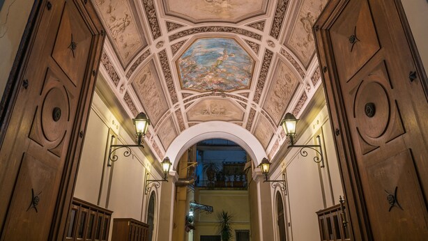 An ornate hallway with a high ceiling, detailed ceiling artwork, and wall-mounted lighting fixtures. - Rome Centro-Repubblica Apartment  (Roma)