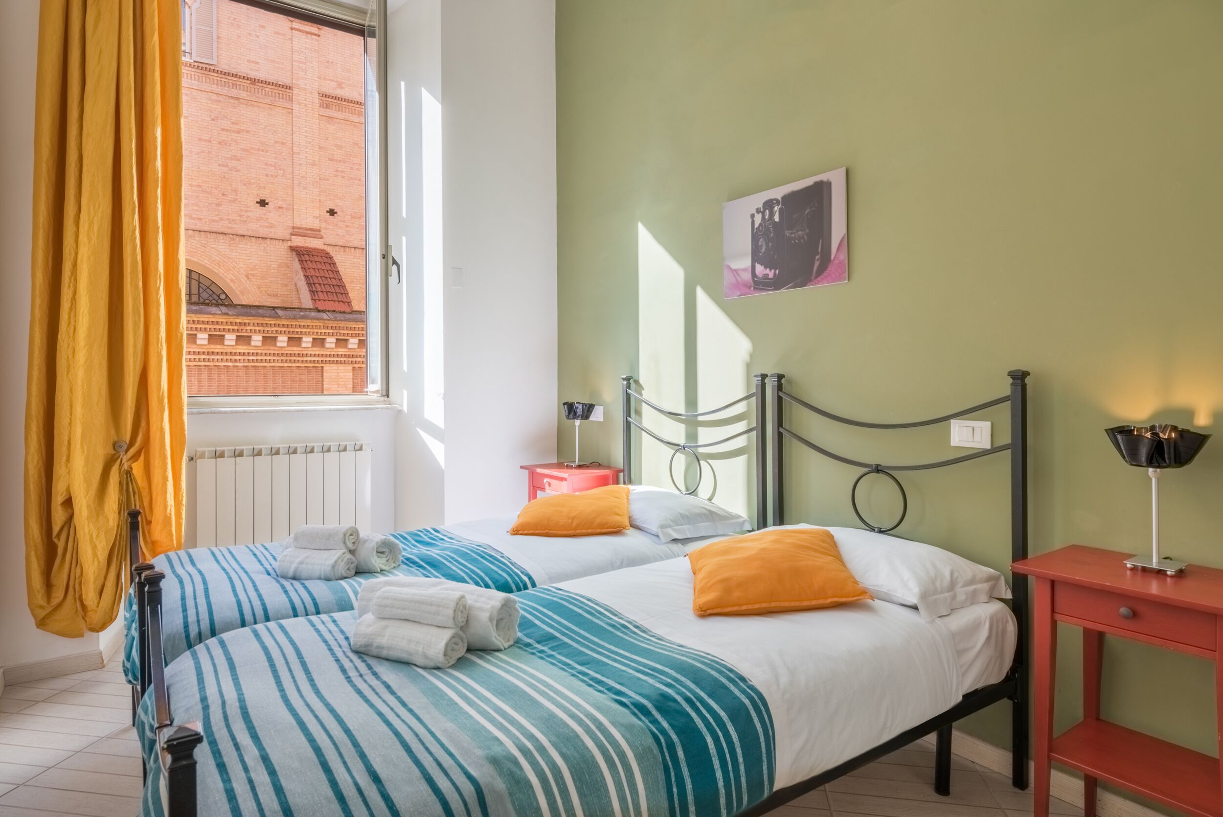 Cama doble con ropa de cama a rayas, una mesita de noche, una lámpara y vistas de un edificio por la ventana.