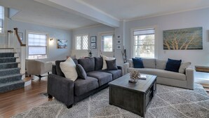 En rummelig stue med en grå sofa, en beige sofa og et sofabord. Der er en trappe til venstre og et stort vindue til højre.