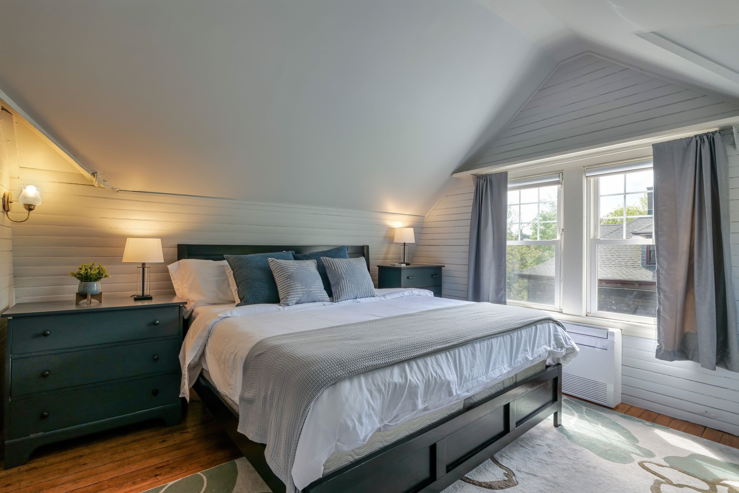 A bedroom with a bed, two bedside tables with lamps, a window with curtains, and a radiator.