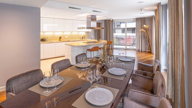 Een moderne eethoek met een lange tafel voor zes personen, een keuken op de achtergrond en grote ramen met gordijnen.