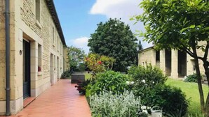 Une cour avec un chemin pavé de carreaux, des bâtiments en pierre et une végétation luxuriante comprenant diverses plantes et arbres.