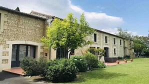 Un bâtiment en pierre avec un toit en tuiles, un petit jardin avec un arbre et des arbustes, et un ciel bleu dégagé.