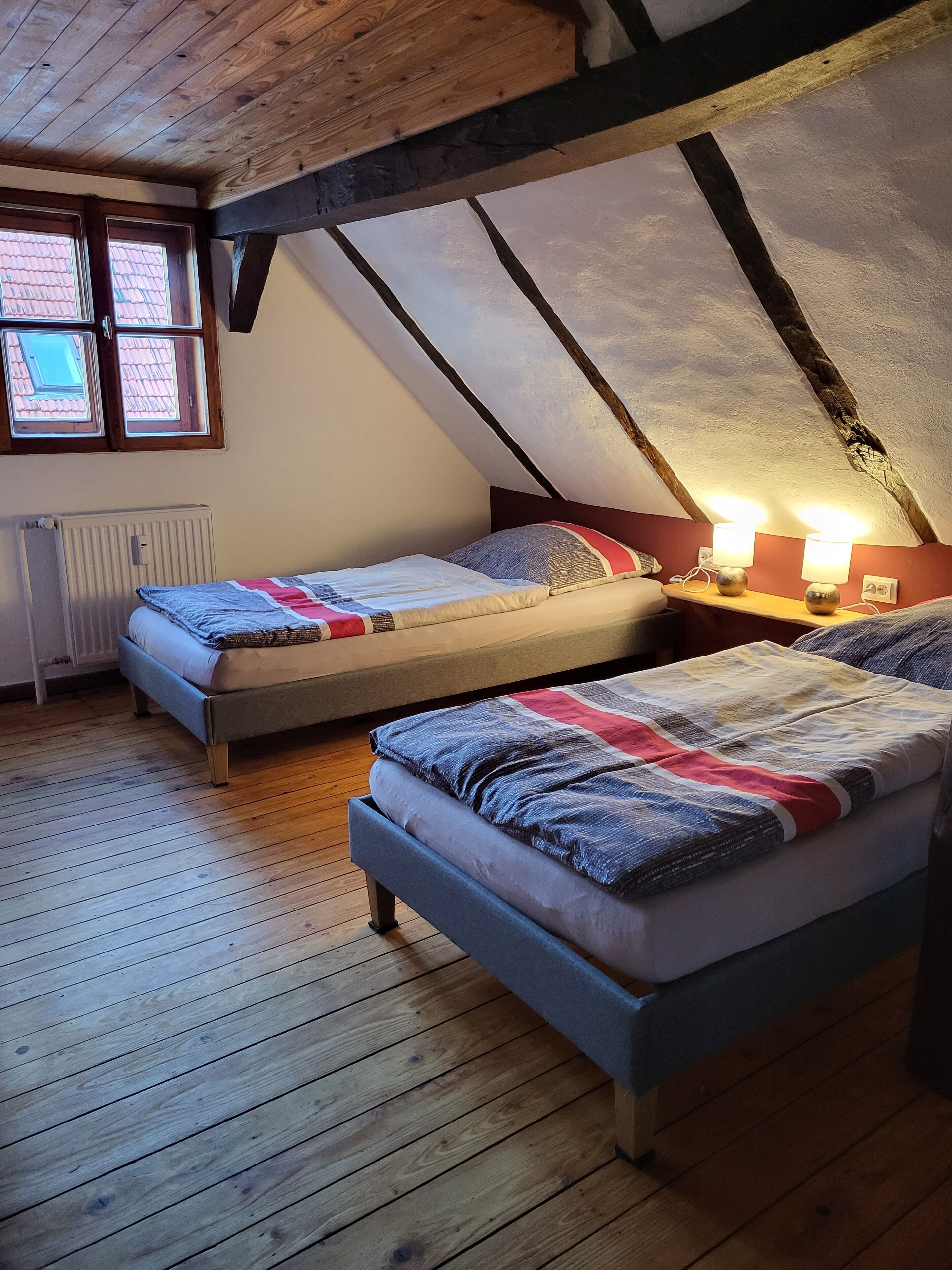 Une chambre avec deux lits, un plancher en bois, un plafond en pente et une fenêtre donnant sur un toit recouvert de tuiles rouges.