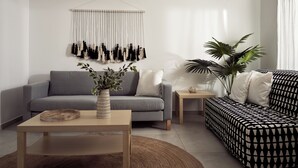 A modern living room with a grey sofa, a patterned ottoman, a small side table, a wooden coffee table, and a macrame wall hanging. - One Bedroom Apartment (Zakynthos)