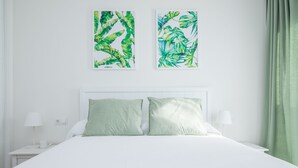 A neatly made bed with two pillows, a white headboard, and two wall-mounted artworks depicting tropical foliage. - SUITE MARINA BEACH COSTA DEL SOL (Fuengirola)