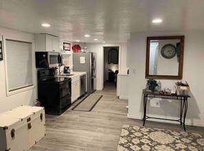 A compact kitchen with a microwave, stove, and refrigerator. There's a small table with a clock and a mirror on the wall. - Cozy 1BR Cottage Near Lake, Trails & City Access (Kenmore)