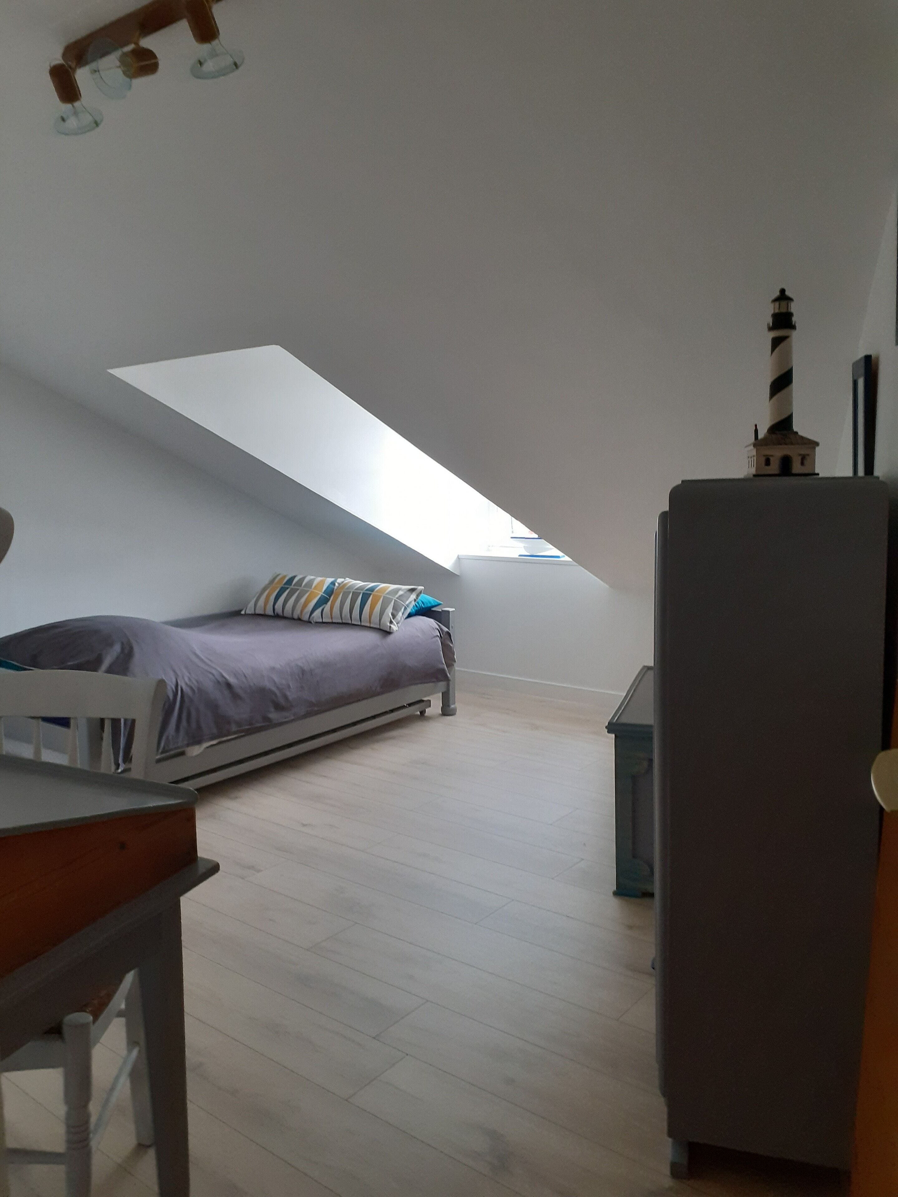 A small, compact room with a sloped ceiling, a bed with a striped pillow, a small table, and a refrigerator.