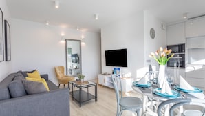 Ein modernes Wohnzimmer mit grauem Sofa, einem Esstisch für vier Personen und einer Küchenzeile mit Flachbildfernseher.