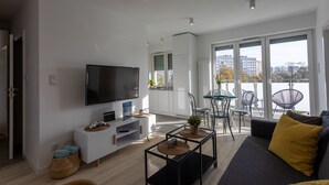 Ein modernes Wohnzimmer mit einem Flachbildfernseher, einer Couch, einer Essecke mit Stühlen und großen Fenstern mit Stadtblick.