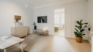 Ein modernes, minimalistisches Zimmer mit Holzboden, einem wandmontierten, weißen Fernseher, einer Holzbank, einem kleinen Tisch und einem Blumentopf.