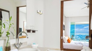 Ett modernt hotellbadrum med en stor tvättställ, ett badkar och utsikt över balkongen med en säng och handdukar.