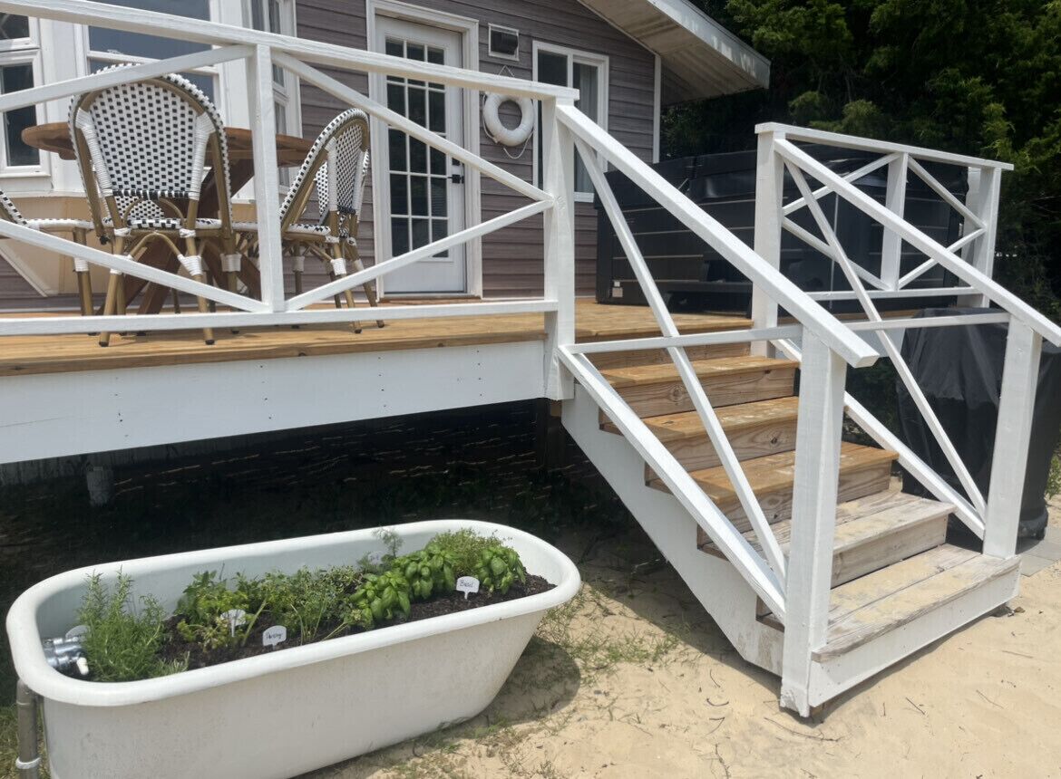 A wooden deck with white railings, a white planter with green plants, and wicker chairs.