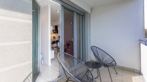Een balkon met twee zwarte stoelen met draadframe en een kleine tafel, een glazen schuifdeur die leidt naar een binnenruimte met een zichtbare keukenhoek.