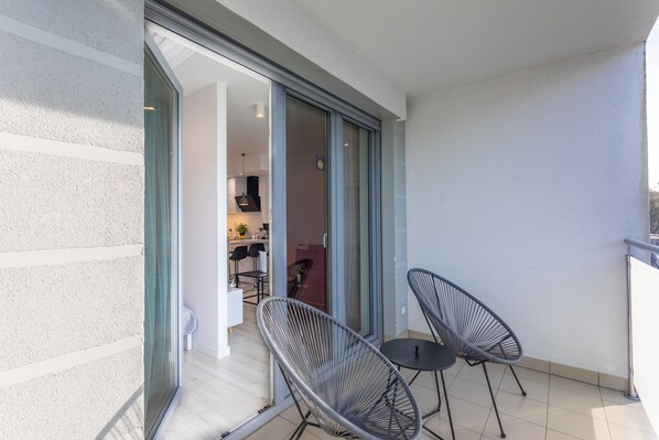 Un balcon avec deux chaises à l’armature en fil de fer noir et une petite table, une porte-fenêtre coulissante donnant sur un espace intérieur où l’on aperçoit un coin cuisine.