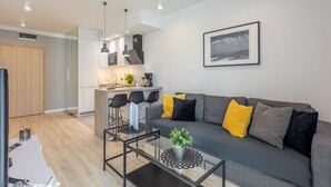 Een moderne woonkamer met een grijze bank, zwarte en gele kussens, een glazen salontafel met een plant, en een keuken op de achtergrond.