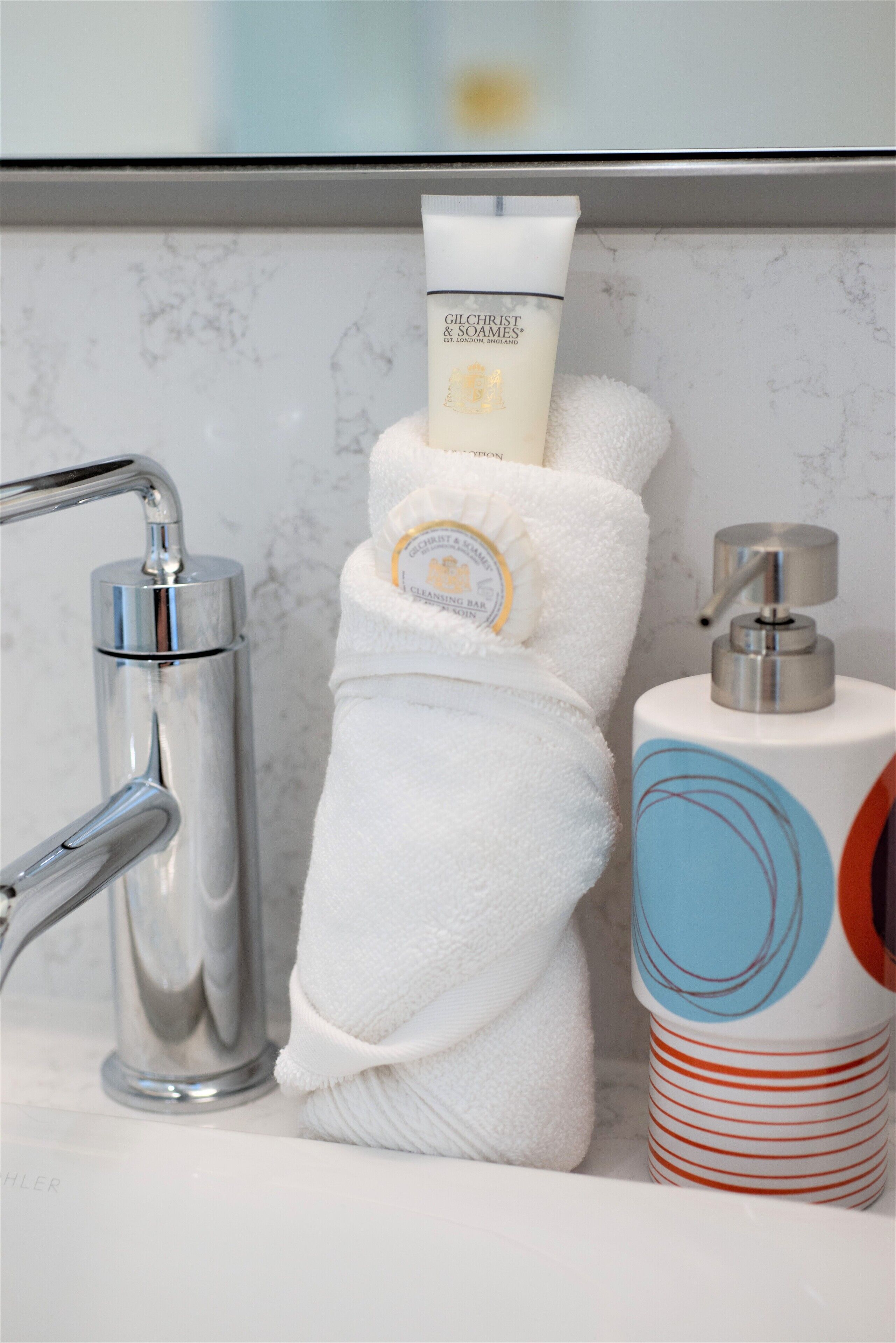 A bathroom with a marble wall, a white towel, a bottle of Gilchrist & Soames body lotion, a soap dispenser, and a striped soap dish.