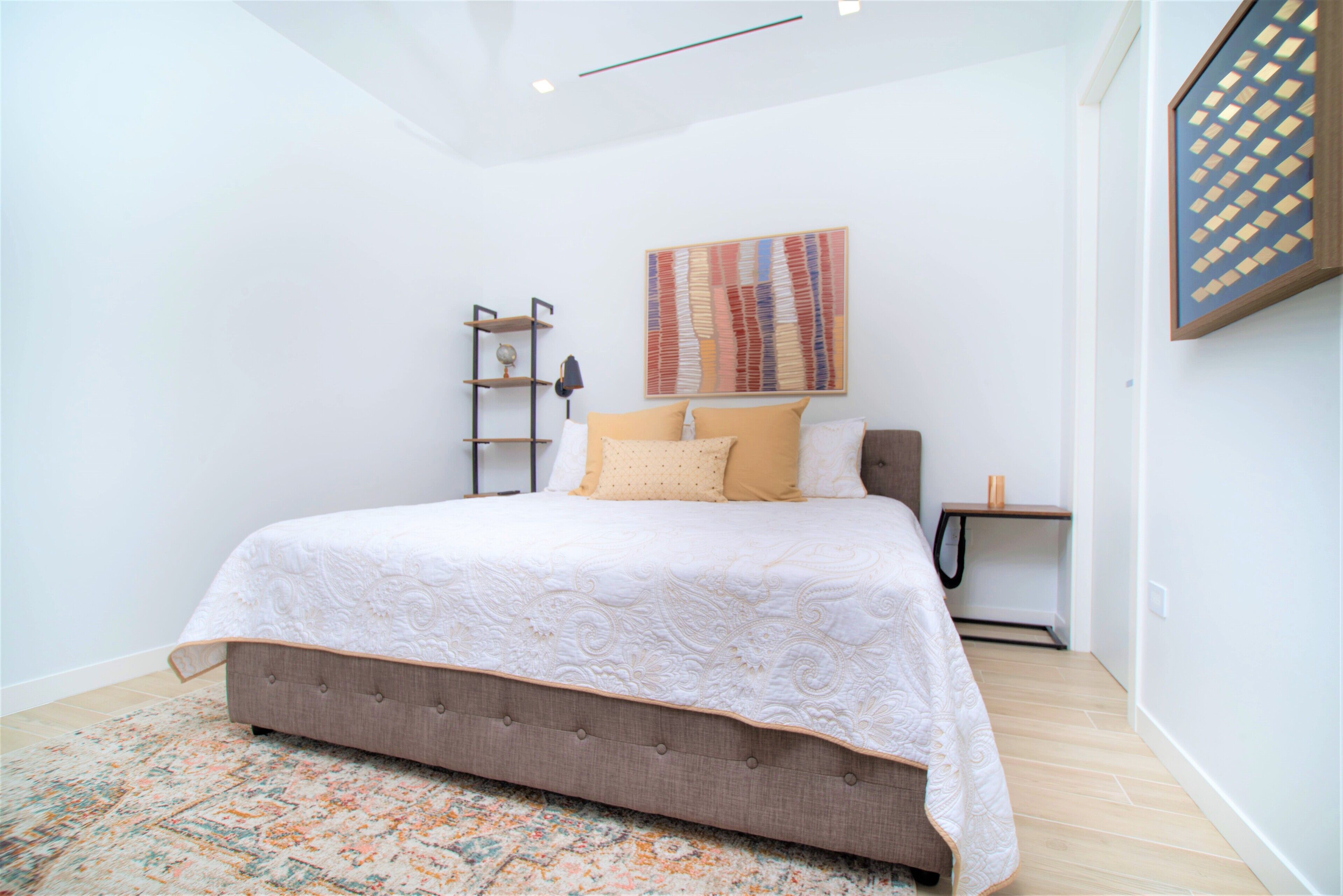 A bedroom with a large bed, a patterned rug, a wall-mounted shelf, and a painting.