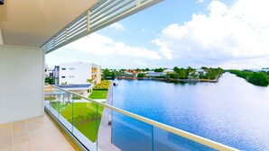 A balcony with a glass railing overlooking a calm river, with buildings and greenery in the background. - Cayman Luxury Rentals at One Canal Point (Seven Mile Beach)